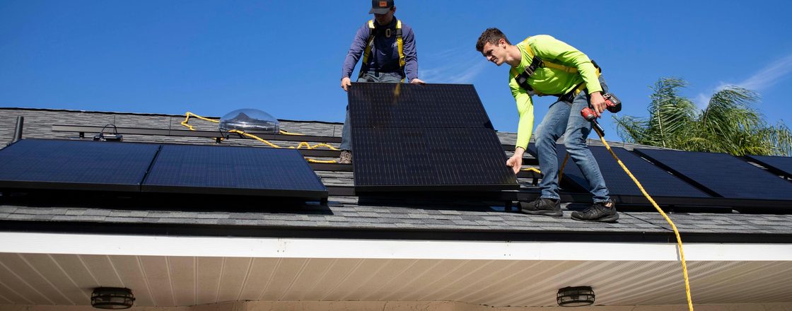 Two installers in safety harnesses fitting solar panels to a shingled residential roof under a clear sky