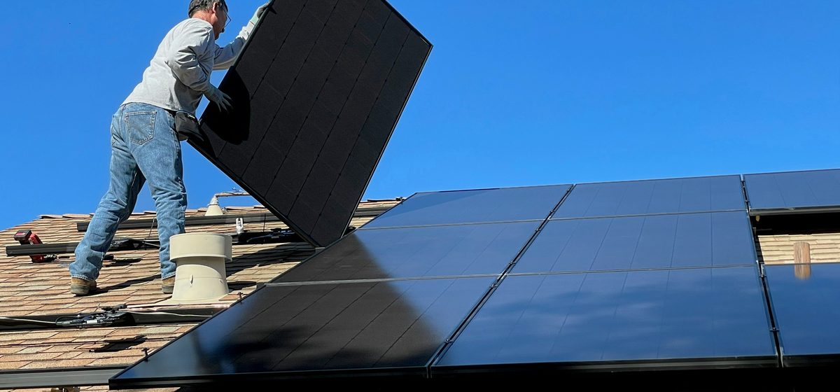 An installer lifting a solar panel into place on a tiled roof beside a completed row of panels