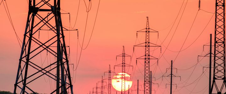 A line of high-voltage transmission towers silhouetted against an orange sunset